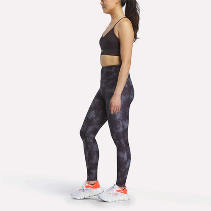A woman stands sideways wearing Reebok Women's High-Rise Printed Leggings, a black sports bra, and white athletic shoes with orange details, against a plain white background.