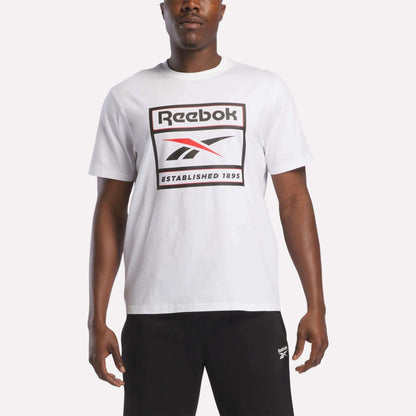 A man stands against a plain background wearing the Reebok Men's Logo T-Shirt in white, featuring the iconic Reebok logo and "Established 1895" printed on the front. Paired with black pants, this soft cotton tee from Reebok is ideal for everyday wear.
