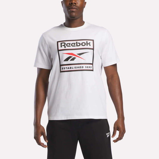 A man stands against a plain background wearing the Reebok Men's Logo T-Shirt in white, featuring the iconic Reebok logo and "Established 1895" printed on the front. Paired with black pants, this soft cotton tee from Reebok is ideal for everyday wear.