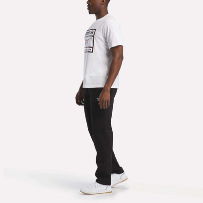 A man stands sideways wearing the Reebok Men's Logo T-Shirt, made from soft cotton fabric, paired with black pants and white sneakers against a plain white background—an ideal outfit for everyday wear.