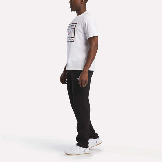 A man stands sideways wearing the Reebok Men's Logo T-Shirt, made from soft cotton fabric, paired with black pants and white sneakers against a plain white background—an ideal outfit for everyday wear.