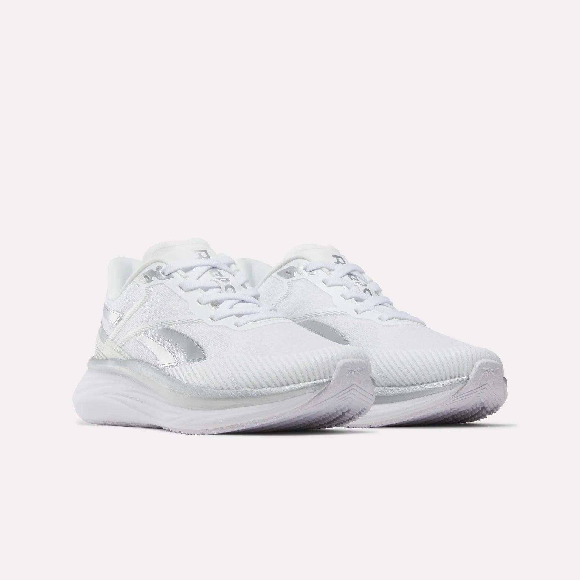 A pair of Women’s Viva Speed Shoes by Reebok in white with silver accents, featuring breathable mesh uppers, a cushioned midsole for enhanced support, thick cushioned soles, and a secure lace-up closure—all showcased against a plain white background.