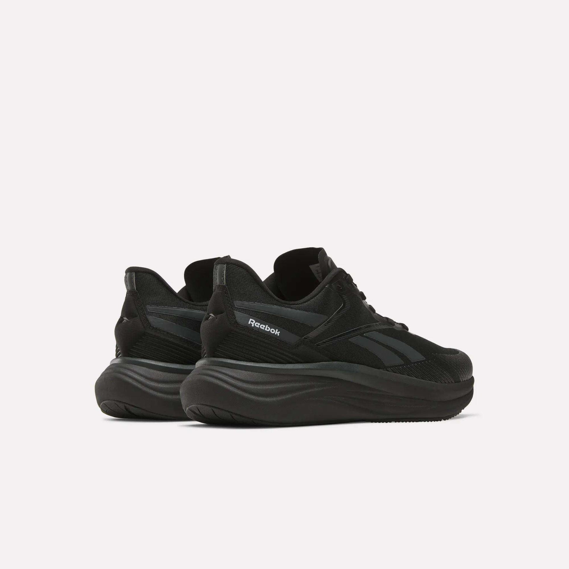 The Reebok Men's Viva Speed Shoes, shown from a rear side angle on a white background, feature black mesh uppers and thick wavy soles, delivering a perfect mix of breathability and cushioned comfort.