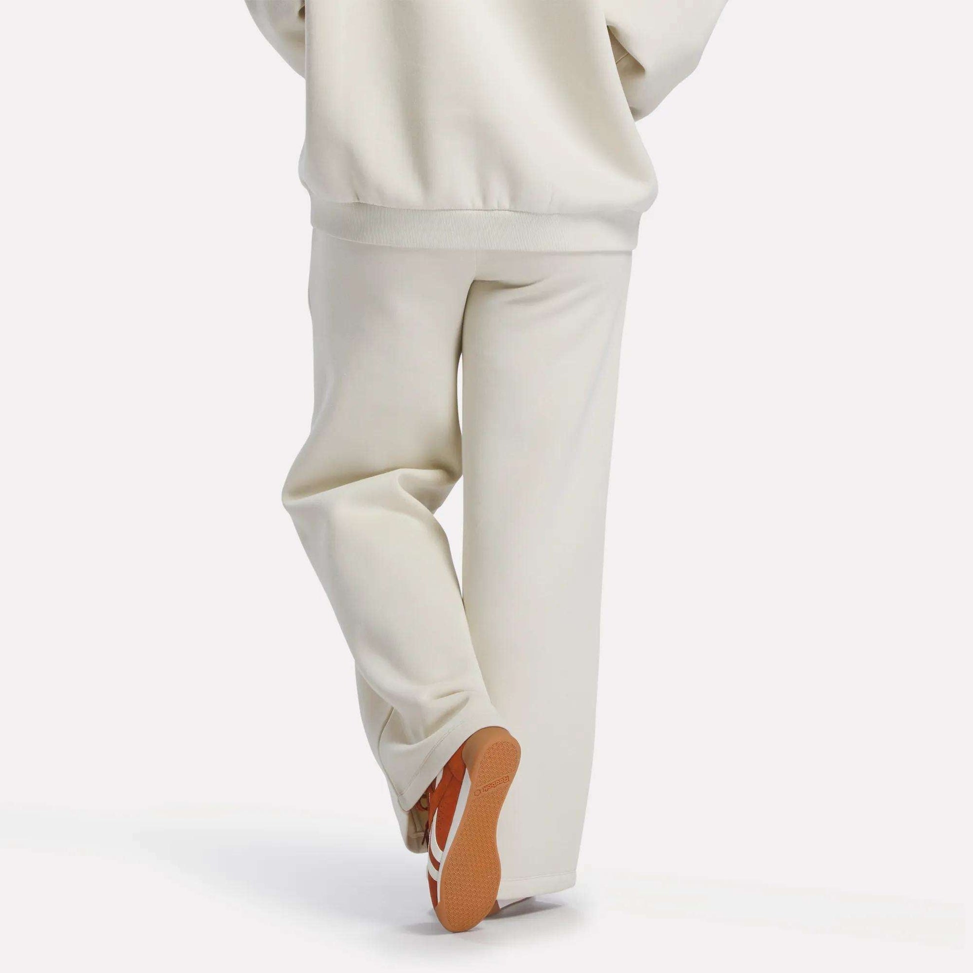 A person in off-white Reebok Women's Heritage Flared Pants and a matching sweatshirt stands with one leg crossed behind the other, revealing the sole of their orange and white sneakers against a plain white background.