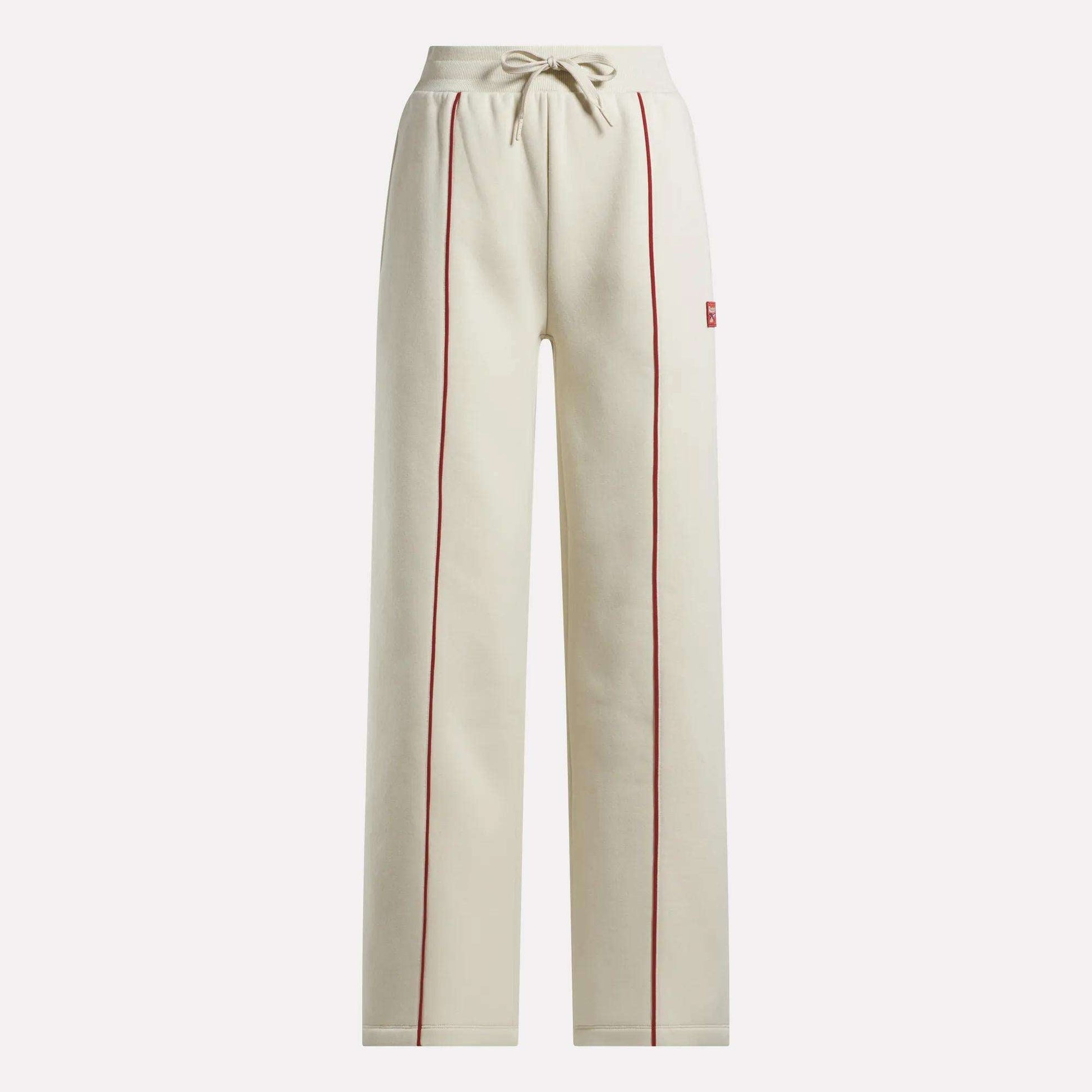 The Reebok Women's Heritage Flared Pants are beige sweatpants made from soft brushed fleece, featuring red piping on the front seams, an elastic drawstring waistband, and side pockets.