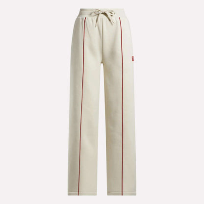 The Reebok Women's Heritage Flared Pants are beige sweatpants made from soft brushed fleece, featuring red piping on the front seams, an elastic drawstring waistband, and side pockets.