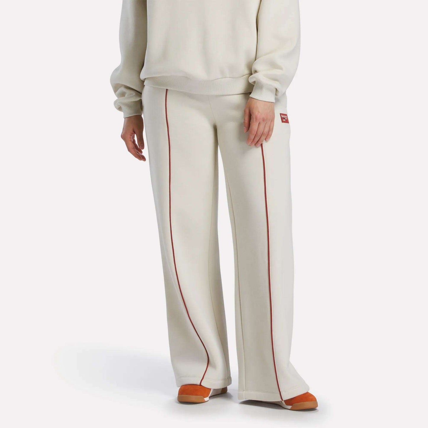 A person wears Reebok Women's Heritage Flared Pants in off-white with red piping, paired with an off-white sweater and orange-beige slippers, creating a sporty vibe against a plain white background.