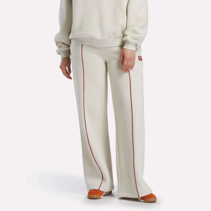 A person wears Reebok Women's Heritage Flared Pants in off-white with red piping, paired with an off-white sweater and orange-beige slippers, creating a sporty vibe against a plain white background.