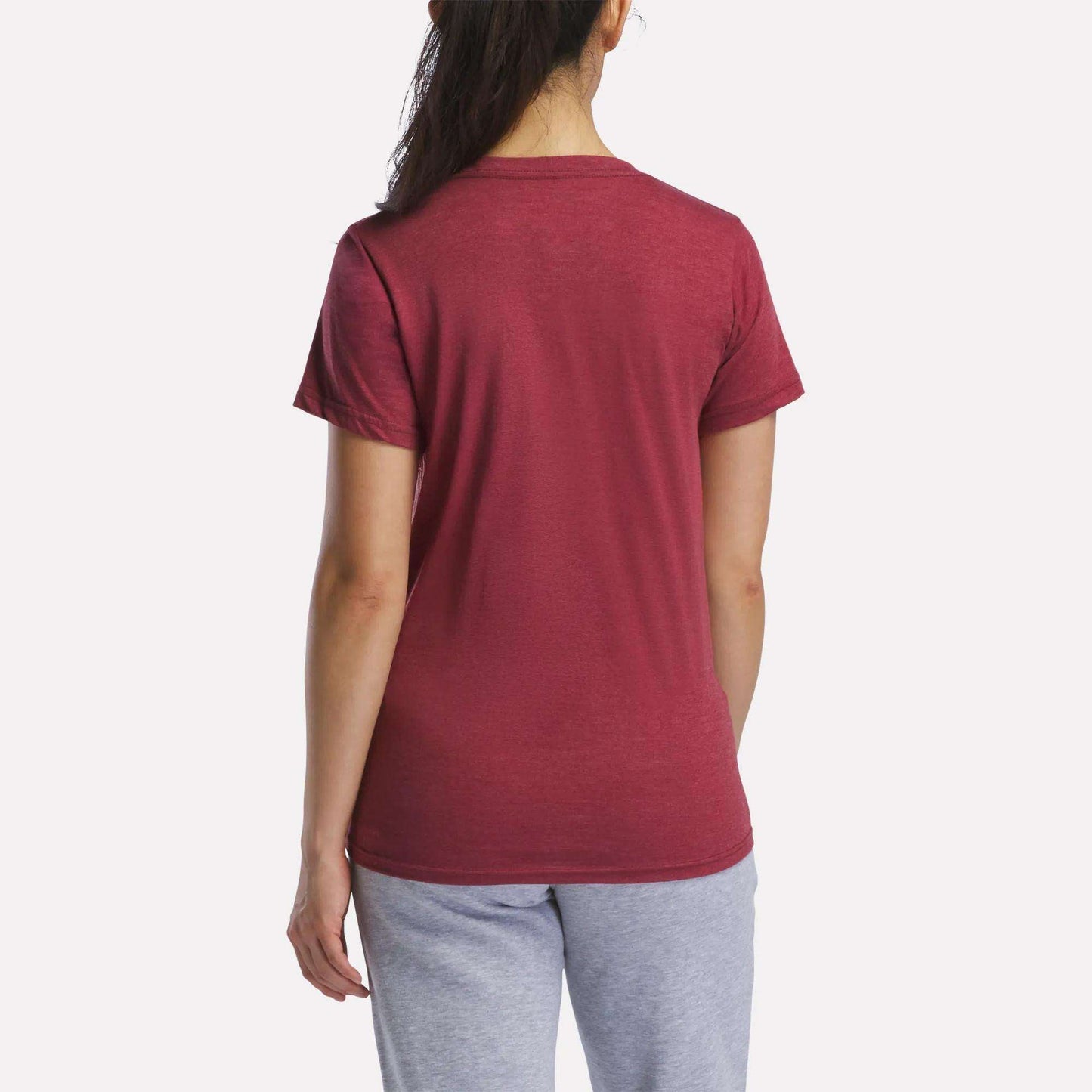 A woman with long dark hair in a ponytail is seen from the back, wearing a Reebok Women's Vector Graphic T-Shirt in maroon with light gray pants, standing against a plain white background.