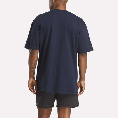 A person stands with their back to the camera, wearing a loose-fit Reebok Men's Stacked Vector T-Shirt in navy blue and black shorts, showing short hair and tattoos on their left arm against a plain white background.
