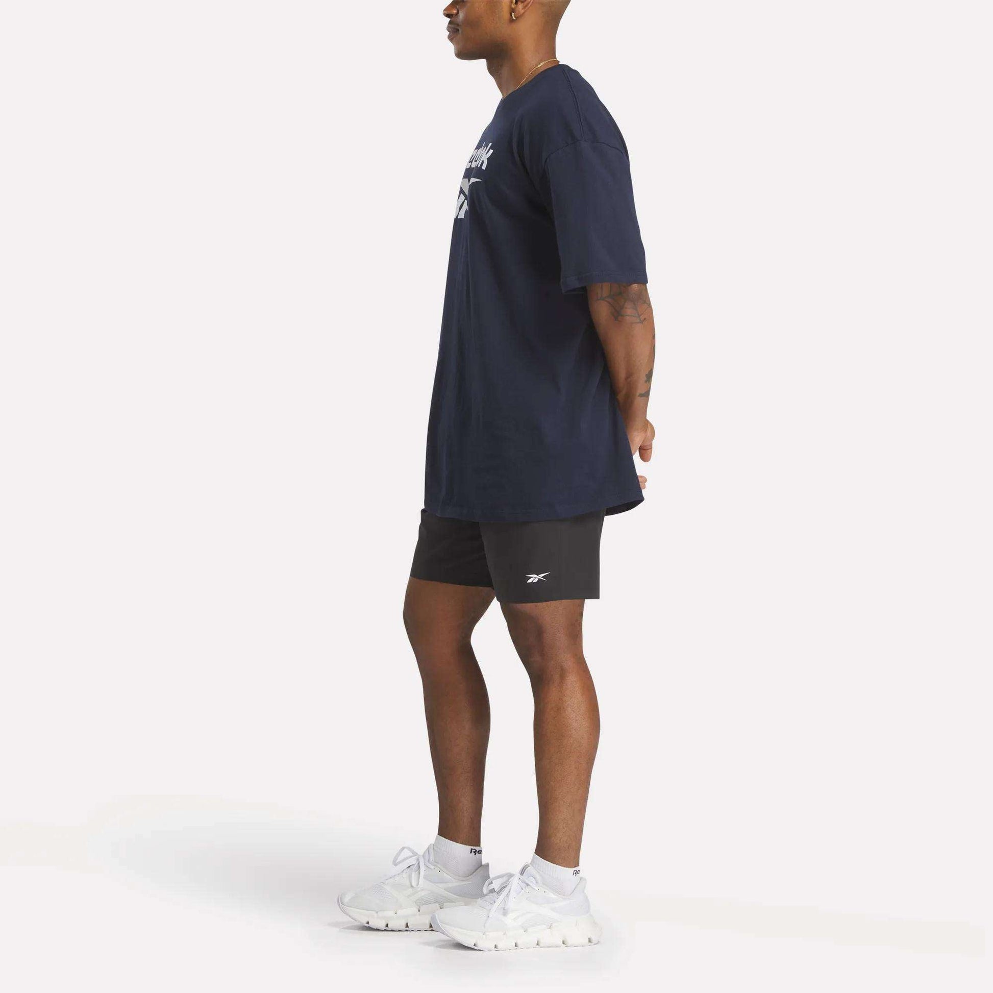 A person stands in profile with hands behind their back, wearing a navy Reebok Men's Stacked Vector T-Shirt, black shorts, white socks, and white Reebok athletic shoes against a plain white background.