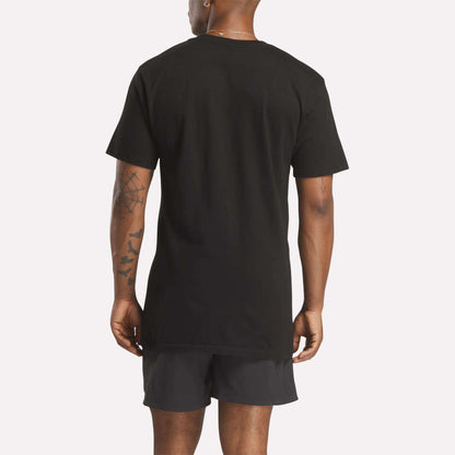 A person stands with their back to the camera, wearing a plain black Reebok Men's Stacked Vector T-Shirt and black shorts. Tattoos are visible on their left arm and both forearms. The background is a solid light color.