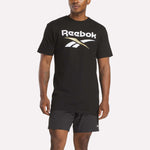 A person models the Reebok Men's Stacked Vector T-Shirt in black, featuring a bold white and gold logo, paired with matching black shorts, set against a plain white background.