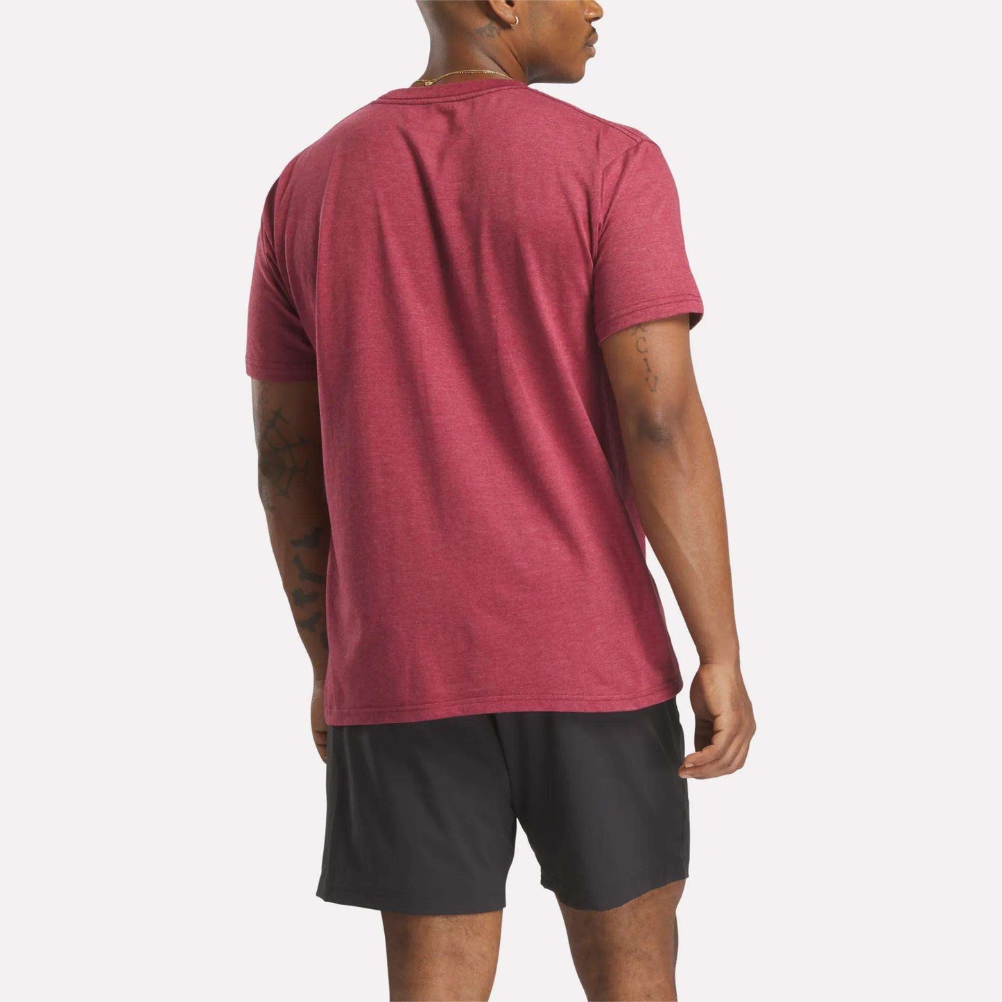 A man faces away from the camera, wearing a red Reebok Men's Stacked Vector T-Shirt and black shorts against a plain white background.