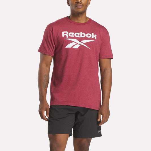 A person models Reebok's Men's Stacked Vector T-Shirt in maroon with a bold white logo, paired with black Reebok shorts, against a plain white background.