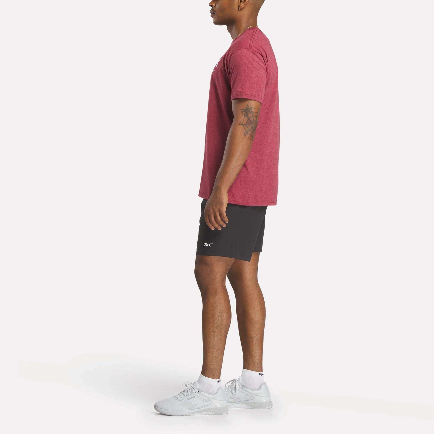 A man in profile wears a red Reebok Men's Stacked Vector T-Shirt, black shorts, white socks, and white athletic shoes against a plain white background.