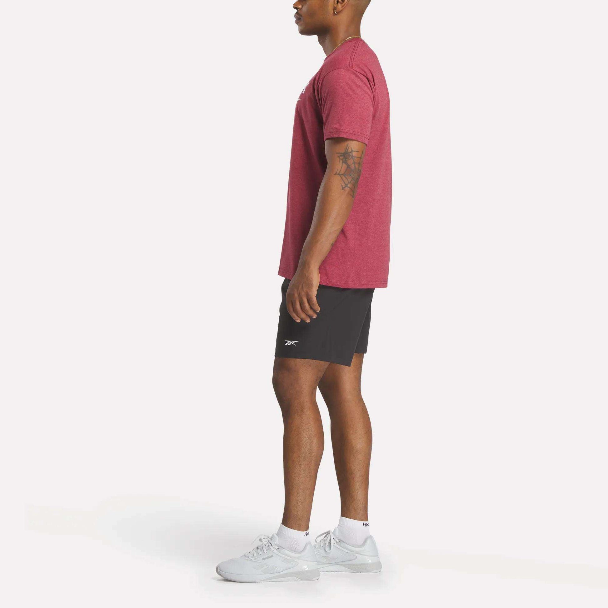 A man in profile wears a red Reebok Men's Stacked Vector T-Shirt, black shorts, white socks, and white athletic shoes against a plain white background.