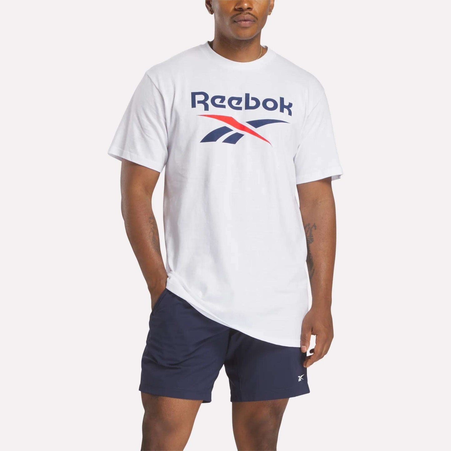 A man wears a white Reebok Men's Stacked Vector T-Shirt featuring a large logo on the chest and navy blue shorts, standing with one hand in his pocket against a plain light background.