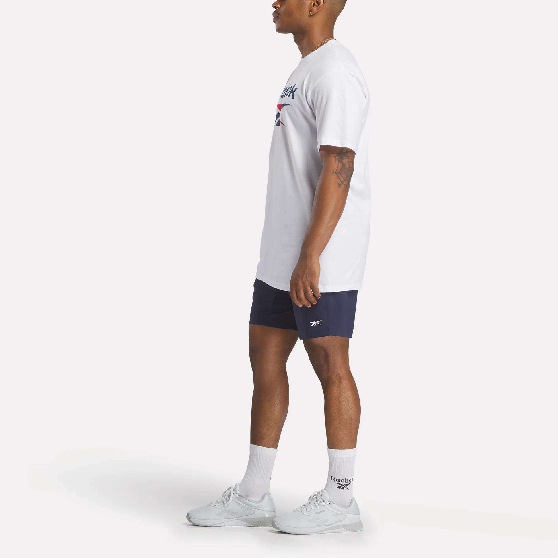 A man stands in profile against a plain light background wearing a Reebok Men's Stacked Vector T-Shirt, navy Reebok shorts, white crew socks, and white sneakers.