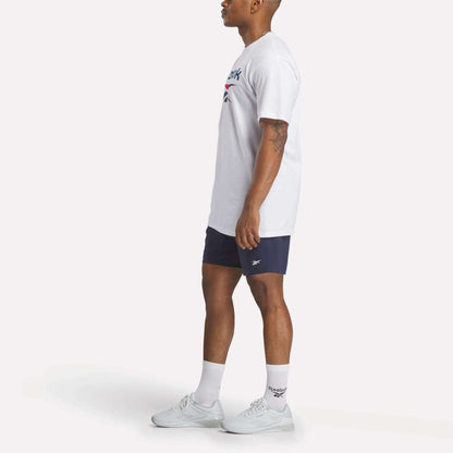 A man stands in profile against a plain light background wearing a Reebok Men's Stacked Vector T-Shirt, navy Reebok shorts, white crew socks, and white sneakers.