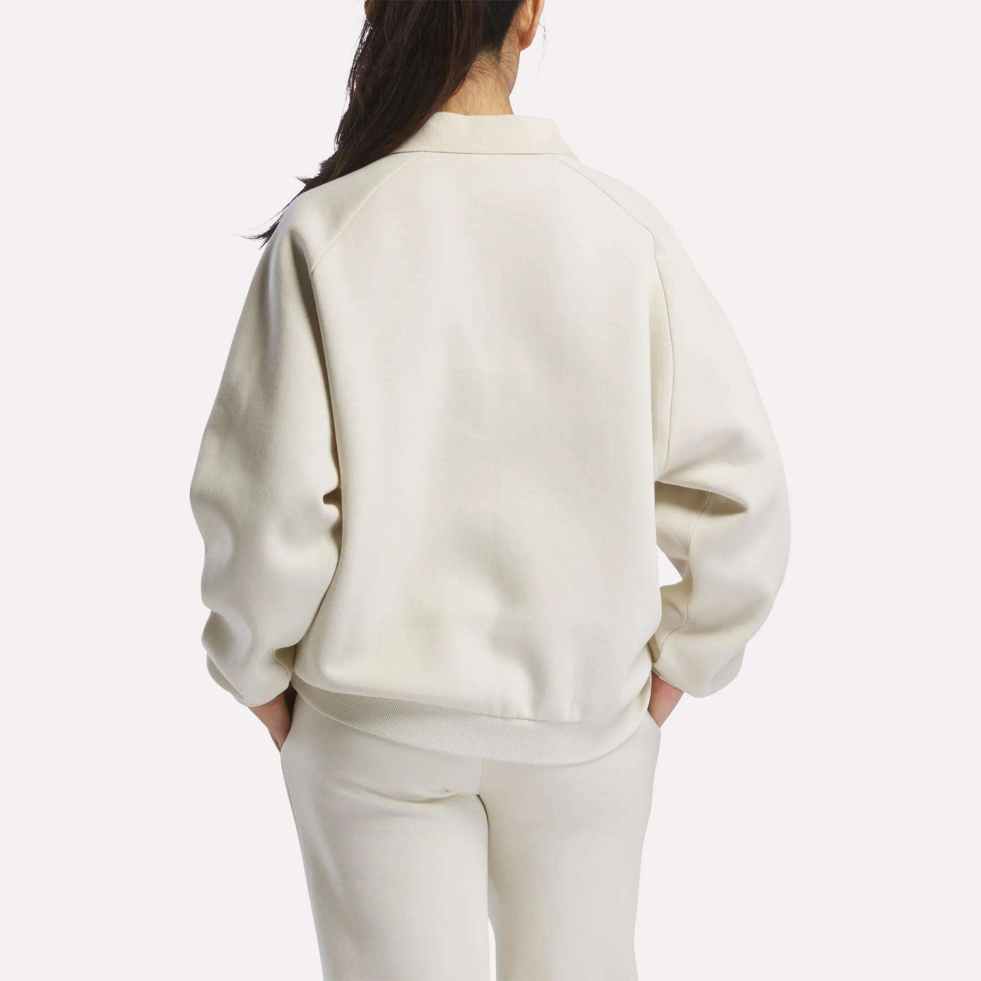 A woman with long dark hair, wearing a cream Women's Heritage Crewneck Sweatshirt by Reebok and matching pants with subtle branding, stands facing away against a plain white background.