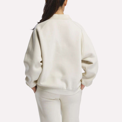 A woman with long dark hair, wearing a cream Women's Heritage Crewneck Sweatshirt by Reebok and matching pants with subtle branding, stands facing away against a plain white background.