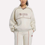 A woman stands with hands in pockets against a white background, wearing the Reebok Women's Heritage Crewneck Sweatshirt and matching pants in cream with brown piping, Sunday League Series text, and cozy brushback fleece.