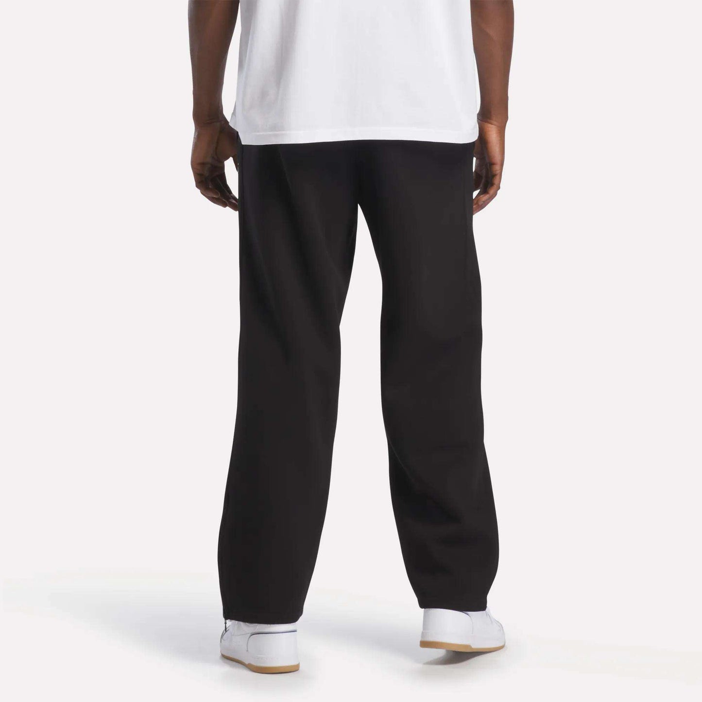 A person is standing facing away, wearing a white T-shirt, Men’s International Straight Leg Pants by Reebok in plush fleece with a relaxed open hem, and white Reebok sneakers. The background is plain and well-lit, showing only the lower part of their body.
