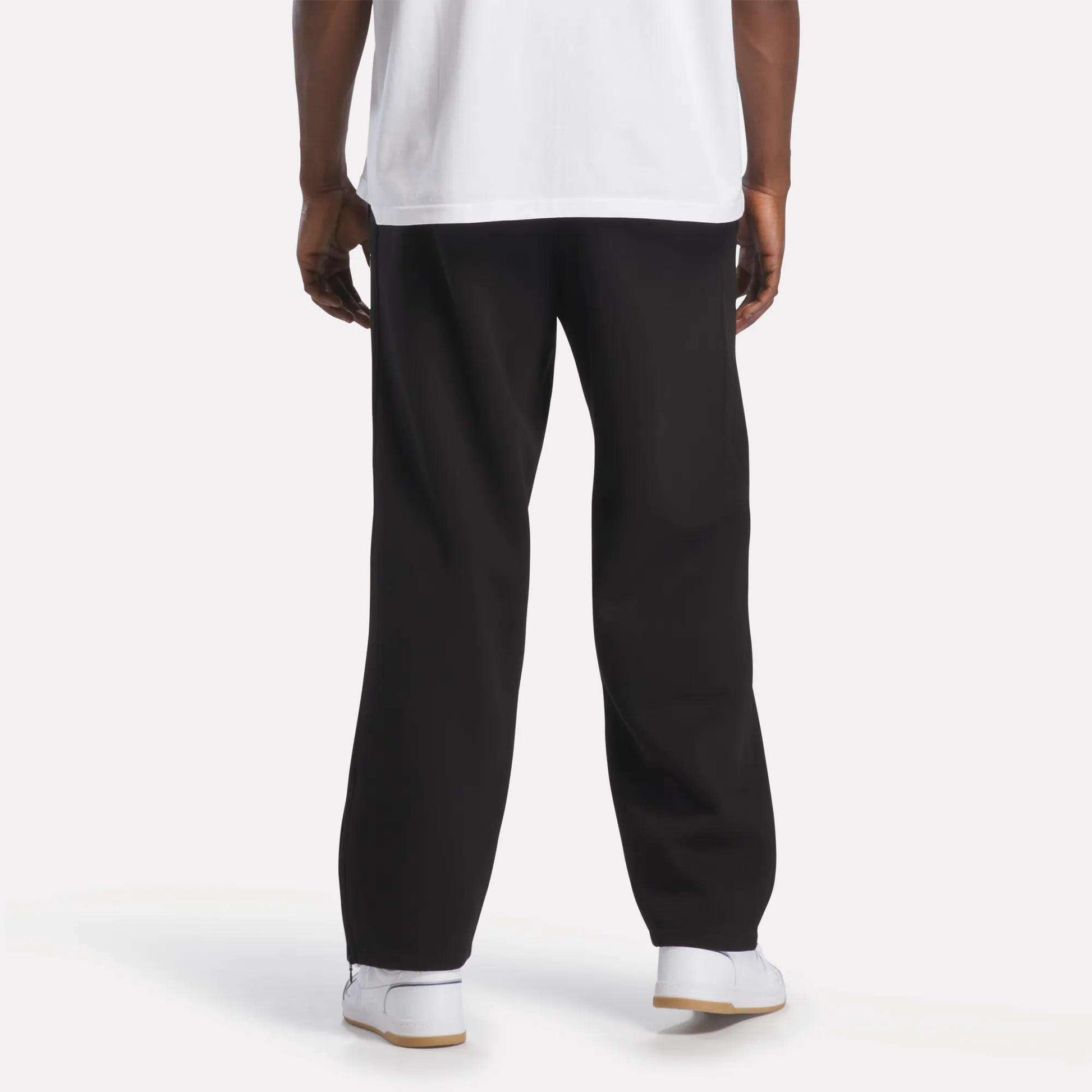 A person is standing facing away, wearing a white T-shirt, Men’s International Straight Leg Pants by Reebok in plush fleece with a relaxed open hem, and white Reebok sneakers. The background is plain and well-lit, showing only the lower part of their body.