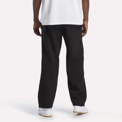 A person is standing facing away, wearing a white T-shirt, Men’s International Straight Leg Pants by Reebok in plush fleece with a relaxed open hem, and white Reebok sneakers. The background is plain and well-lit, showing only the lower part of their body.