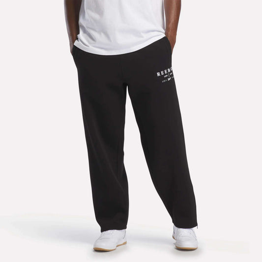 A person wearing black Reebok Men's International Straight Leg Pants, a white T-shirt, and white sneakers stands with hands in pockets against a plain background.