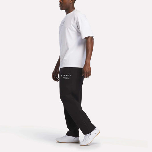 A man wearing a white t-shirt, white sneakers, and black Reebok Men's International Straight Leg Pants stands against a plain white background, looking to the side.