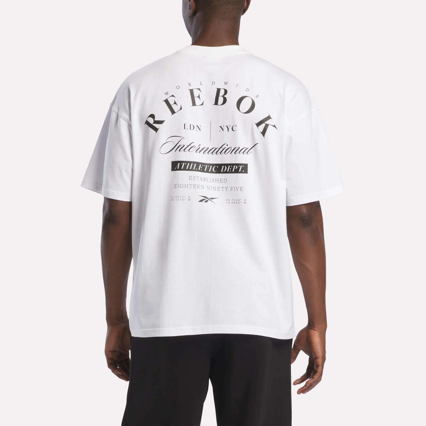 A man models the Reebok Men's International T-Shirt, made from soft white cotton jersey, featuring bold black text and logo on the back—displaying "Reebok International" alongside various city names—as he faces away from the camera.