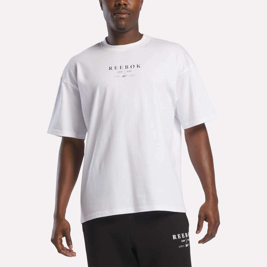 A man stands against a plain white background wearing the Reebok Men's International T-Shirt in white, crafted from soft cotton jersey with black text, paired with black Reebok shorts.