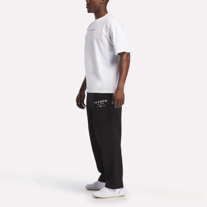 A man stands sideways against a plain white background, wearing the Reebok Men's International T-Shirt made from soft cotton jersey, paired with black Reebok sweatpants and white sneakers.