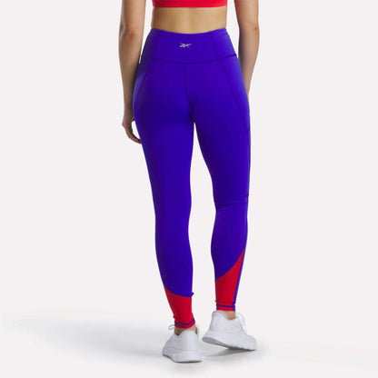 A person wearing a red sports bra, white sneakers, and Reebok Women's Running Leggings—high-waisted purple leggings featuring mesh panels and bold red accents near the ankles—stands with their back to the camera.