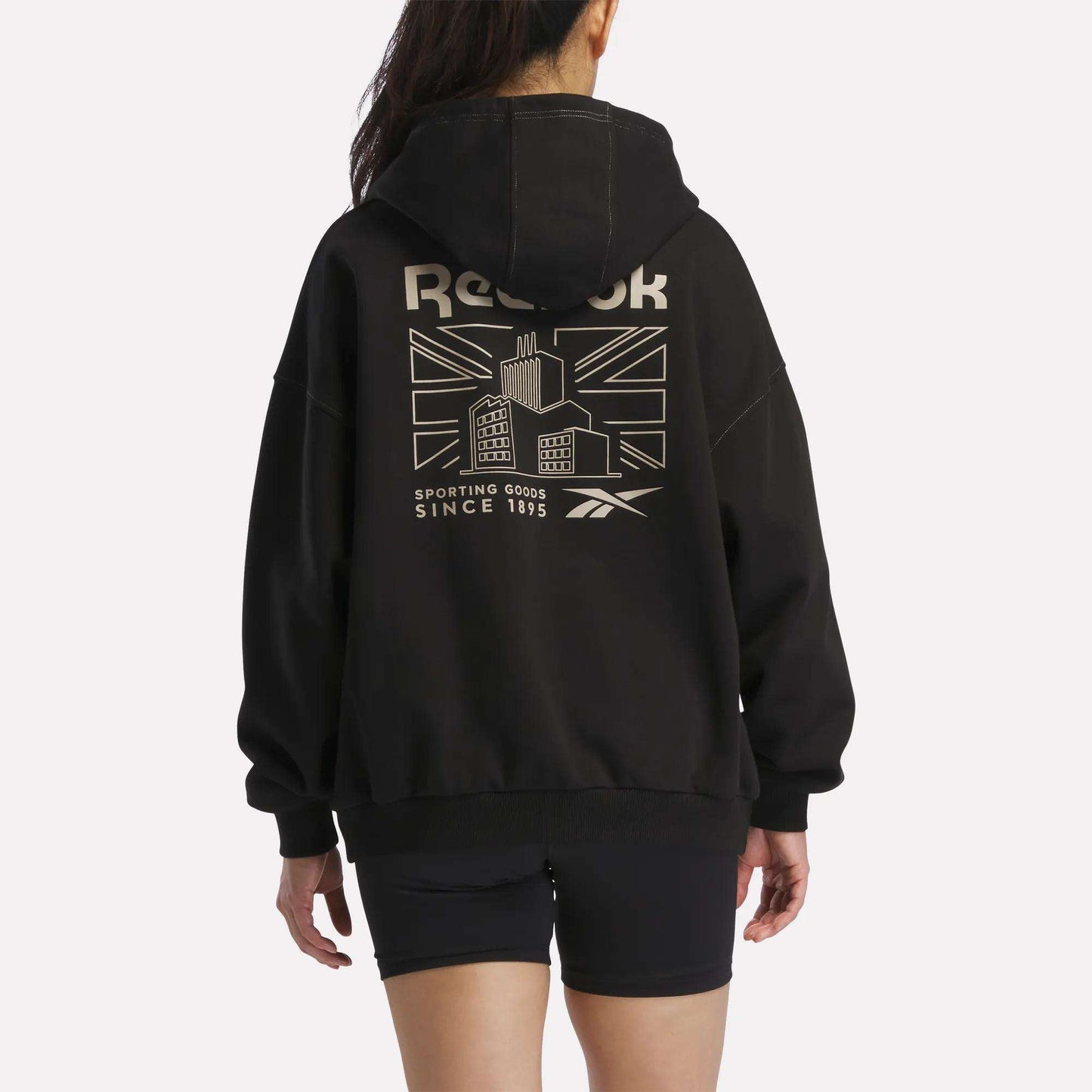 A person is seen from behind wearing the Reebok Women's Relaxed Fleece Hoodie in black, which showcases a bold Union Jack, a building graphic, and text on the back. Paired with black shorts, this fleece hoodie combines comfort and standout style with its striking design elements.