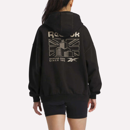 A person is seen from behind wearing the Reebok Women's Relaxed Fleece Hoodie in black, which showcases a bold Union Jack, a building graphic, and text on the back. Paired with black shorts, this fleece hoodie combines comfort and standout style with its striking design elements.