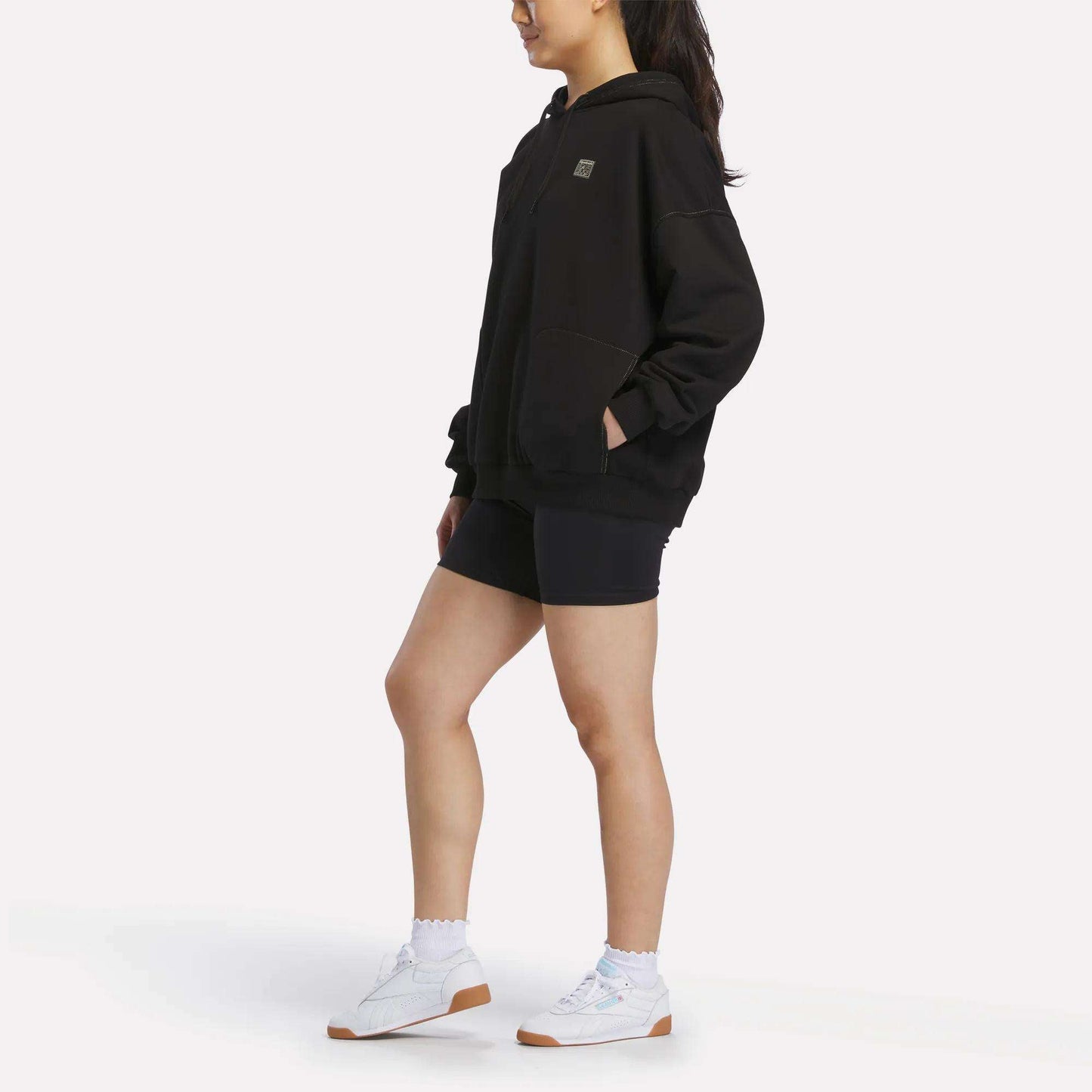 A person stands sideways against a plain white background wearing the Reebok Women's Relaxed Fleece Hoodie in black, paired with black shorts, white socks, and white sneakers.