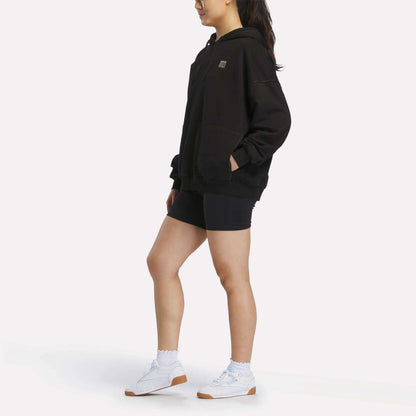 A person stands sideways against a plain white background wearing the Reebok Women's Relaxed Fleece Hoodie in black, paired with black shorts, white socks, and white sneakers.