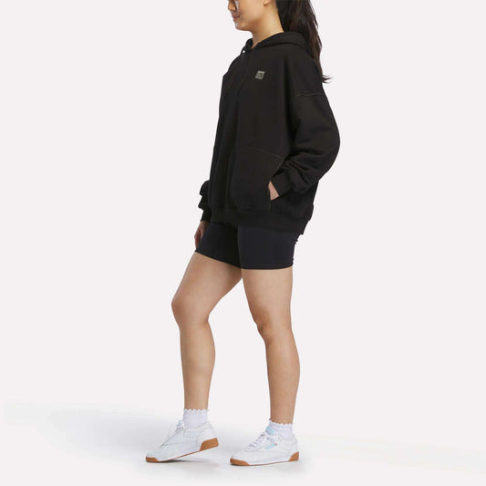 A person stands sideways against a plain white background wearing the Reebok Women's Relaxed Fleece Hoodie in black, paired with black shorts, white socks, and white sneakers.