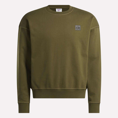The Reebok Men's Patch Logo Crewneck Sweatshirt features a relaxed fit in olive green with long sleeves, ribbed cuffs and hem, crew neck, and a small square logo patch on the left chest. Displayed against a plain light background.