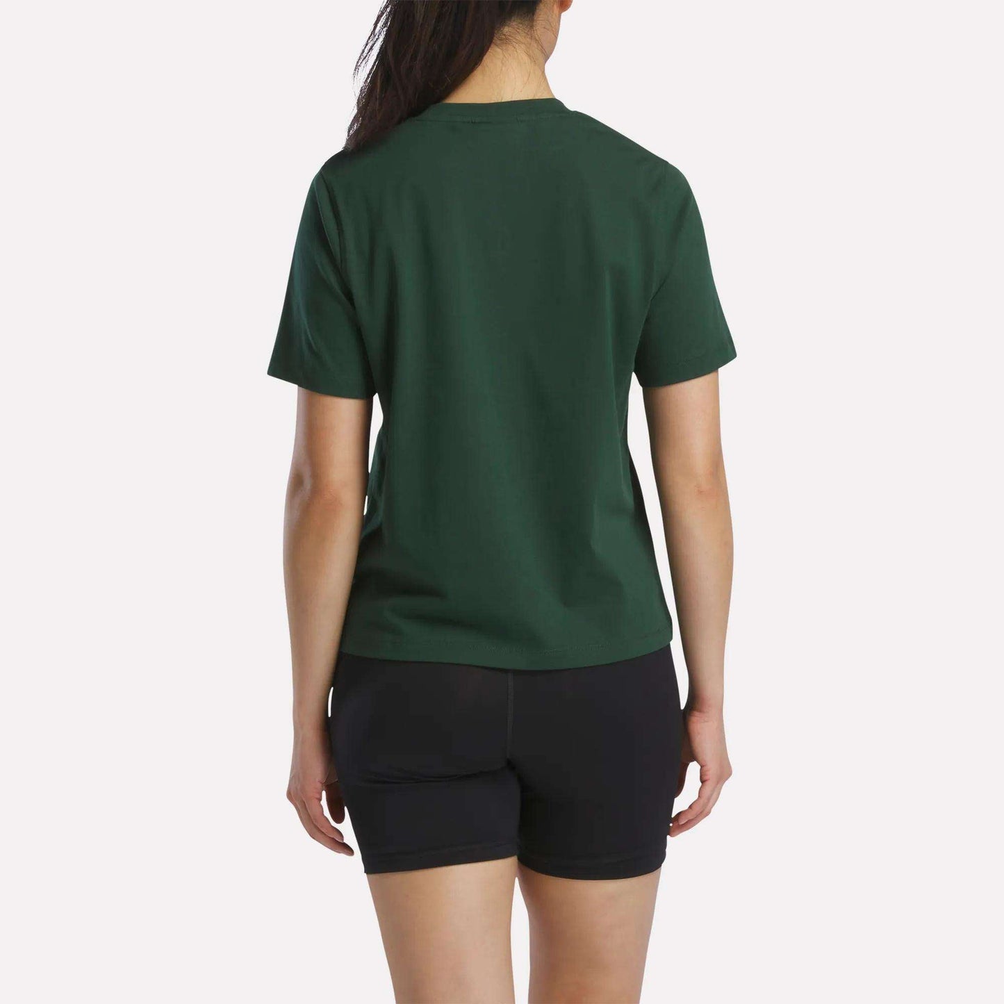 A person stands facing away, wearing a Women's Fitted Graphic T-Shirt by Reebok in dark green and black shorts, set against a plain light background.