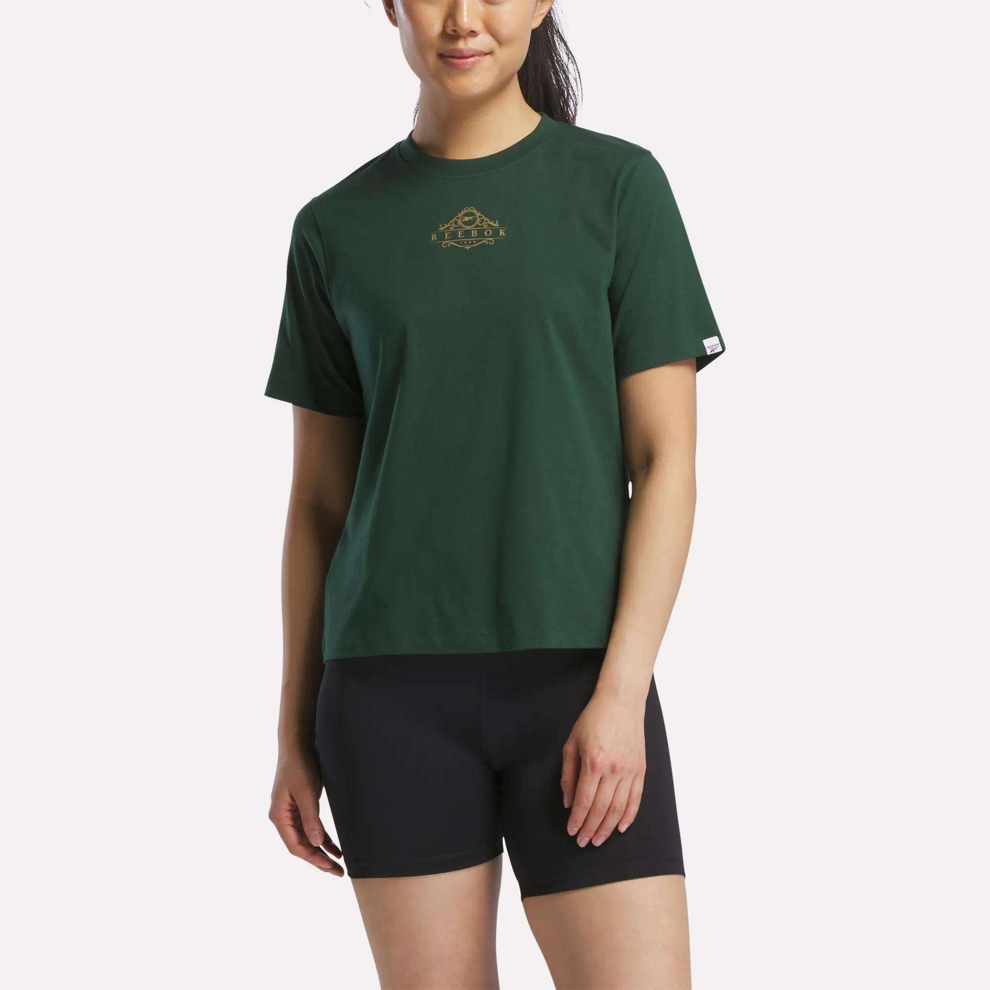 A woman wears the Reebok Women's Fitted Graphic T-Shirt in Heritage Green, featuring a small gold graphic on the chest, paired with black shorts and standing against a plain light background. Crafted from soft cotton, this tee offers a blend of comfort and style for an effortlessly casual look.