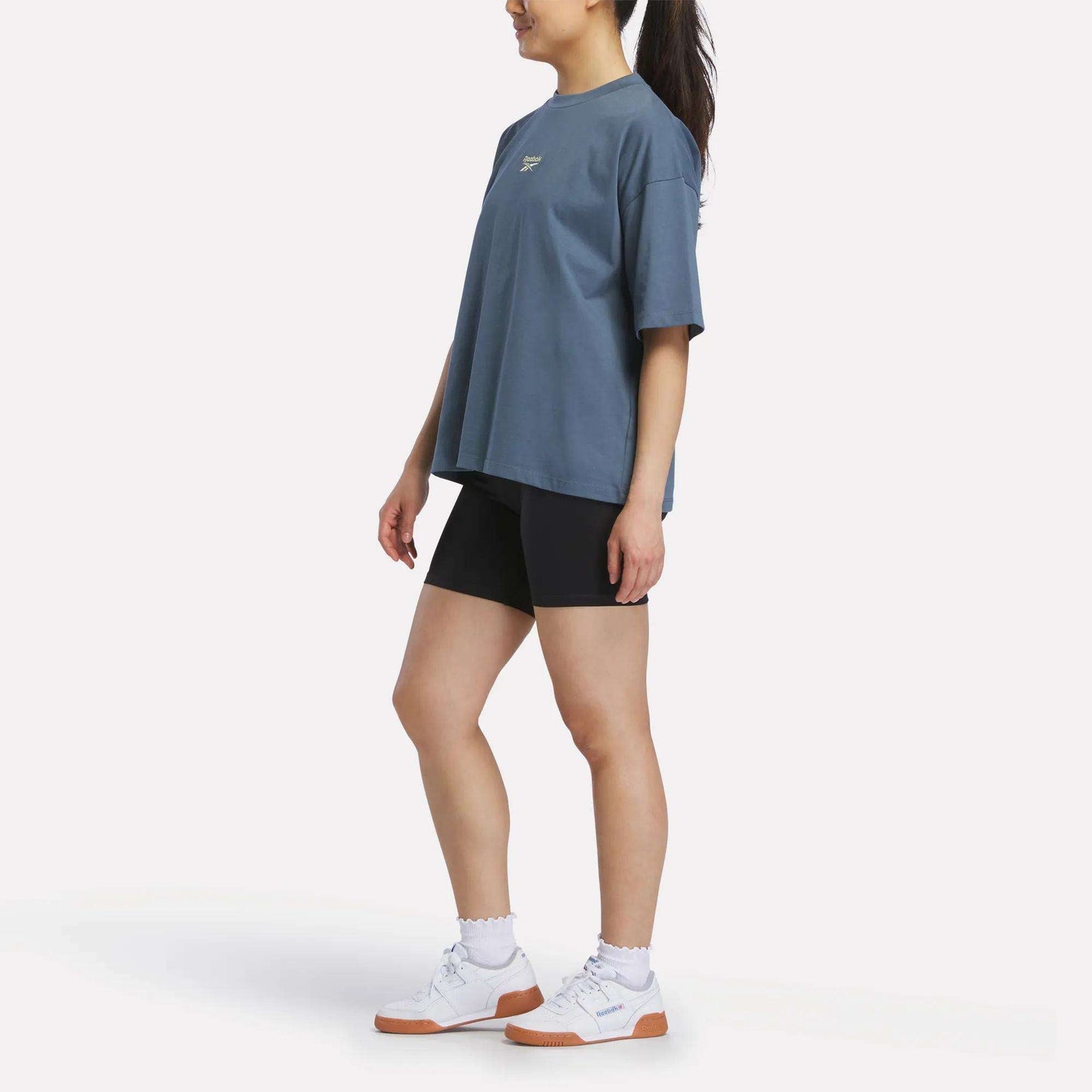 A person stands sideways against a plain white background wearing a Reebok Women's Boxy T-Shirt in soft cotton jersey, paired with black shorts, white socks, and white sneakers with brown soles.
