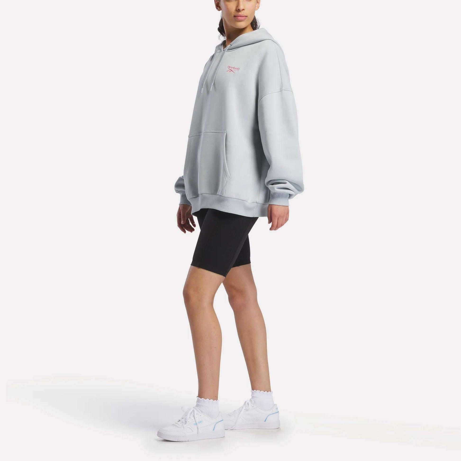 A person wearing a Reebok Women's Relaxed Fleece Hoodie in light gray, styled with black bike shorts, white socks, and white sneakers stands against a plain white background.