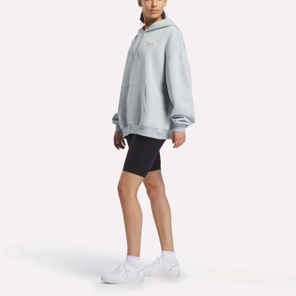 A person wearing a Reebok Women's Relaxed Fleece Hoodie in light gray, styled with black bike shorts, white socks, and white sneakers stands against a plain white background.