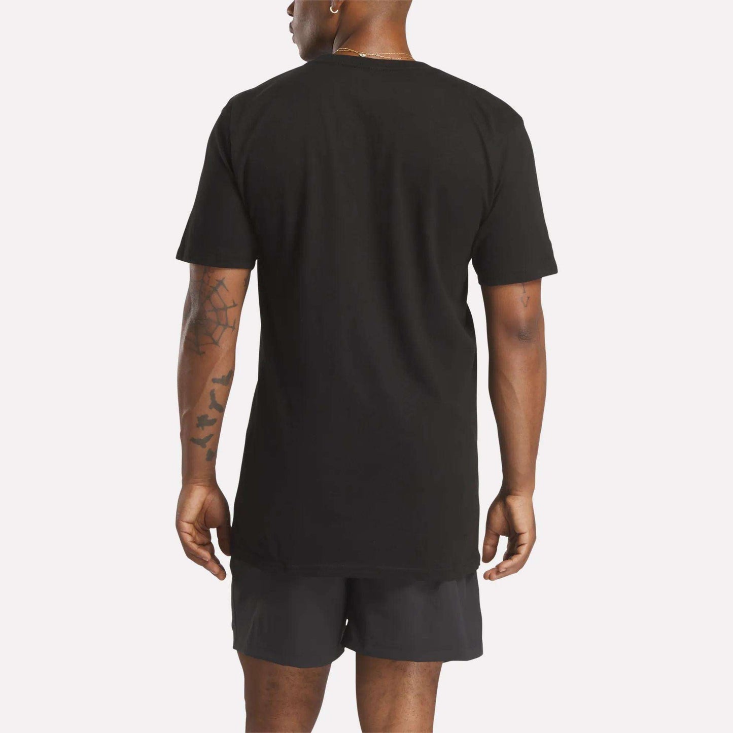 A person with tattoos on their left arm stands facing away from the camera in a relaxed fit Reebok Men's Identity Small Logo T-Shirt and black shorts. The background is plain and light-colored, showcasing the back of the outfit.