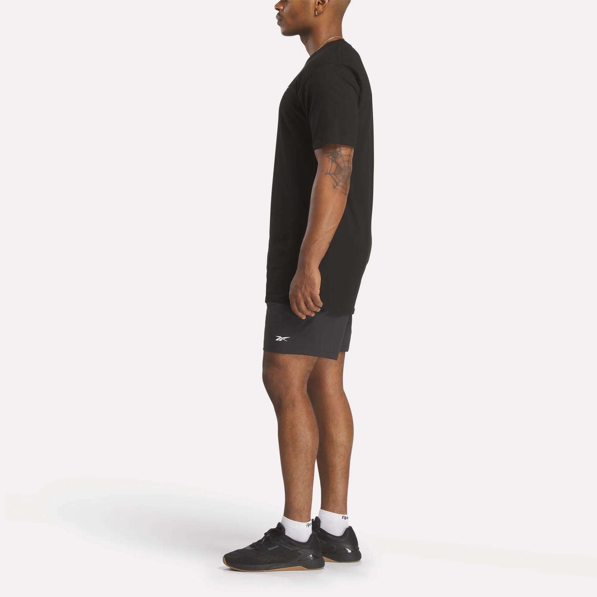 A person in profile wears a relaxed fit black Reebok Men's Identity Small Logo T-Shirt, dark athletic shorts, white socks, and black Reebok sneakers against a plain white background.