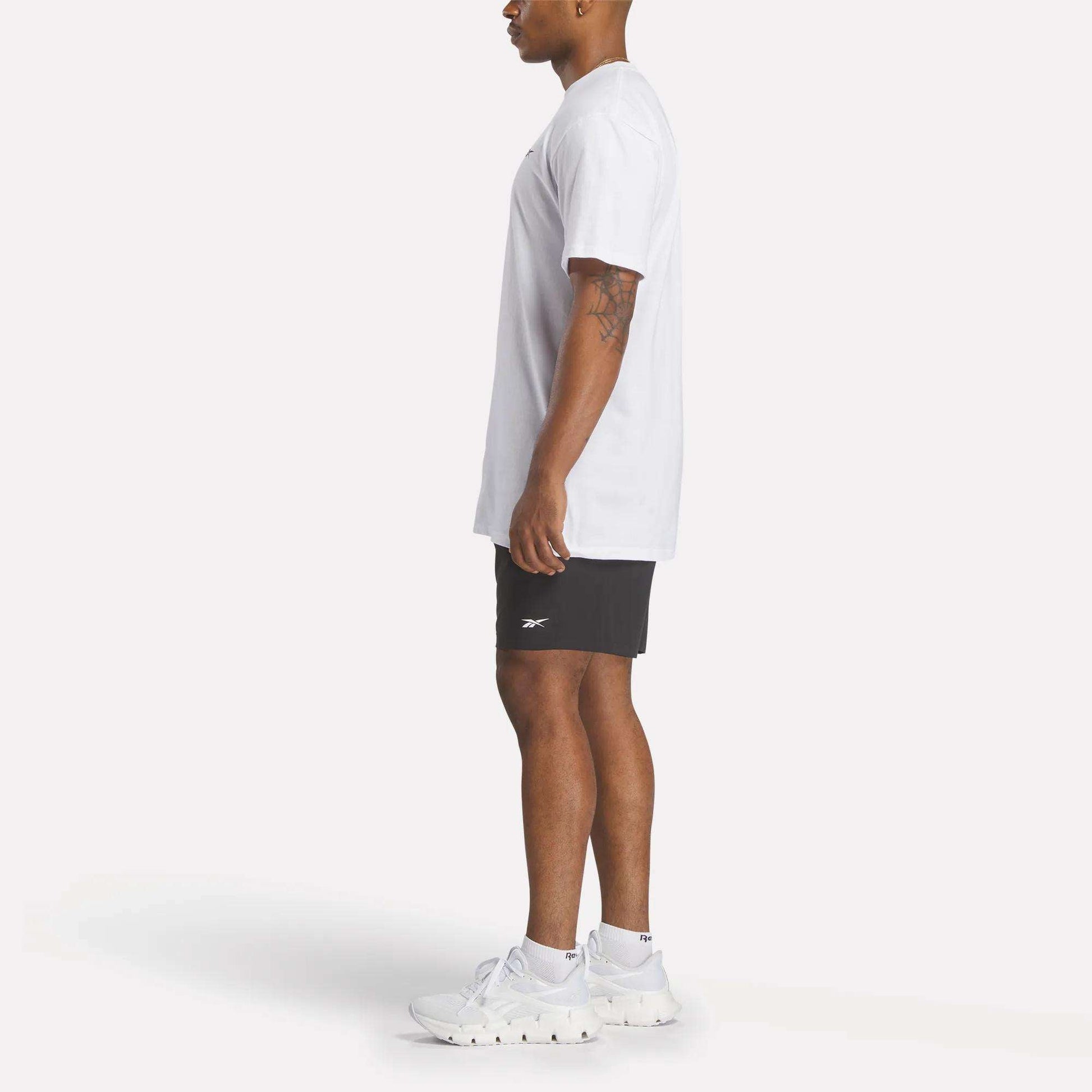 A man stands in profile against a white background wearing a relaxed fit Reebok Men's Identity Small Logo T-Shirt, black Reebok shorts, and white athletic shoes.